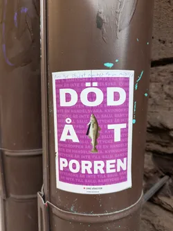 The sticker, adhered to a brown pipe, features bold white text on a purple background, reading "DÖD ÅT PORREN." The background is filled with repeated text in purple. A small fish is illustrated hanging from a noose in the middle of the word "AT."