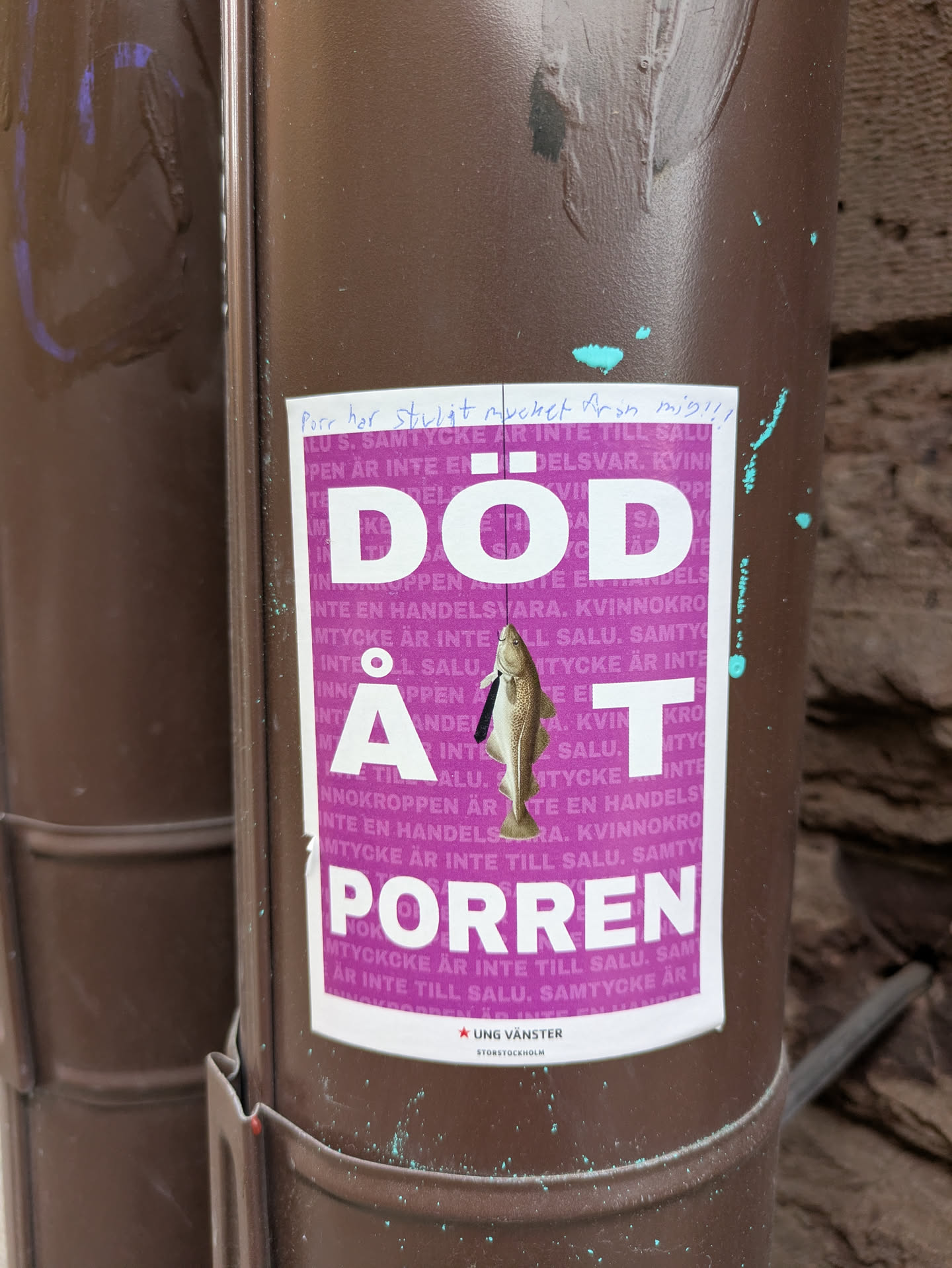 The sticker, adhered to a brown pipe, features bold white text on a purple background, reading "DÖD ÅT PORREN." The background is filled with repeated text in purple. A small fish is illustrated hanging from a noose in the middle of the word "AT."