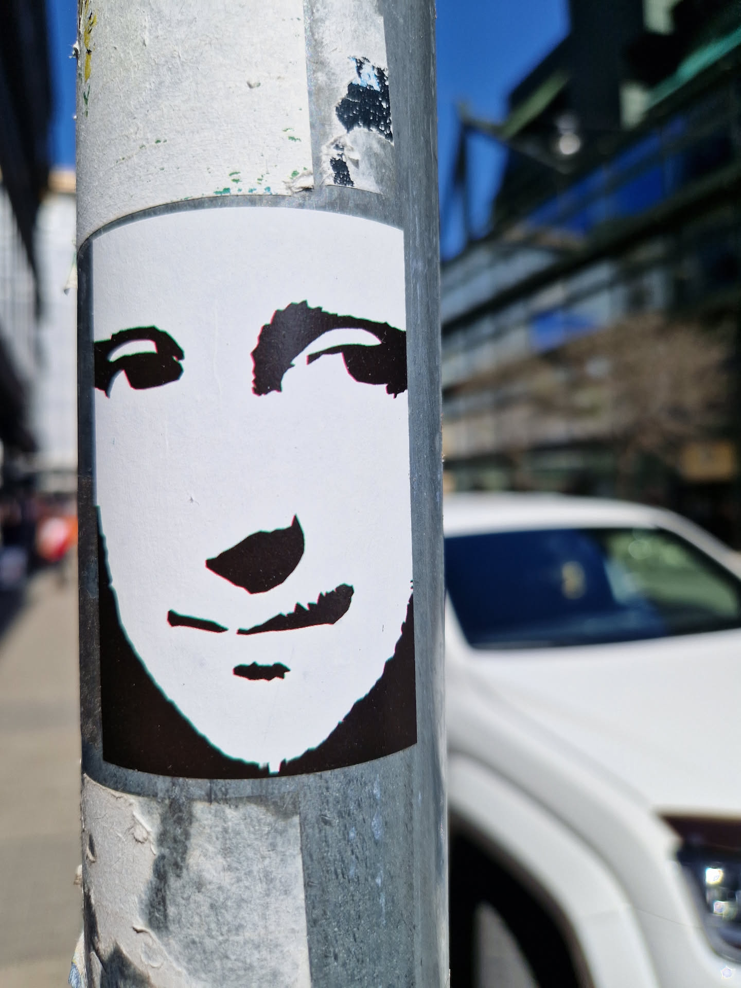A black and white sticker depicting a stylized face is affixed to a metal pole. The sticker is rectangular and features a minimalist design, emphasizing the eyes and mouth. The background is white, and the face is rendered in black with a slight grunge effect around its edges, suggesting it may have been torn or weathered. The surrounding environment is blurred but appears to be an urban setting.