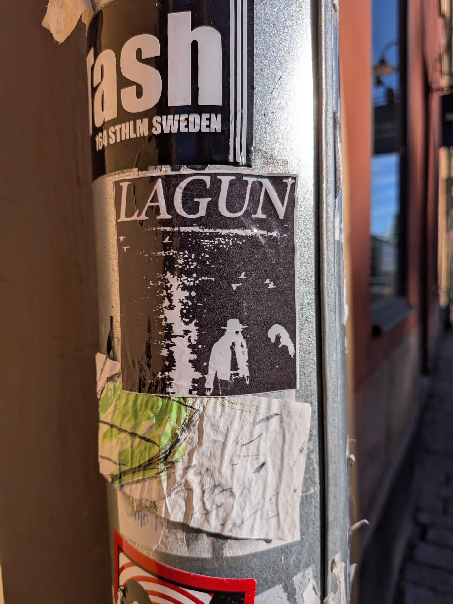 A rectangular black-and-white sticker affixed vertically to a cylindrical metal post. The top section contains the word 'LAGUN' in bold, white serif lettering with a dark outline. Beneath the text is a high-contrast, grain-textured image showing a body of water reflecting light, small birds flying in the sky, and silhouettes of figures—one notably wearing a wide-brimmed hat—standing in the lower right foreground.
