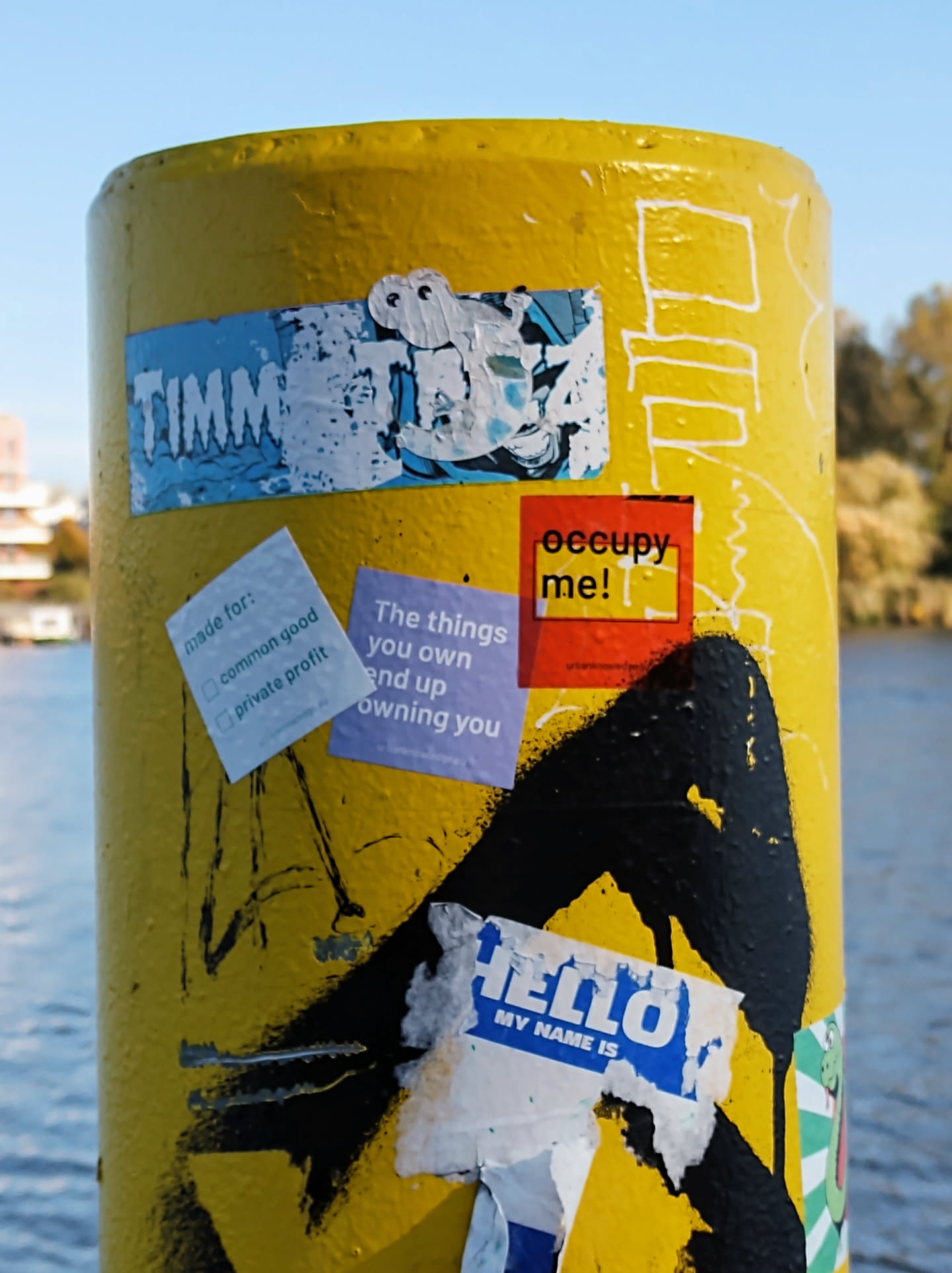 Close-up view of a yellow pole with various stickers attached.  The most prominent sticker features the word "TIMM" in stylized lettering. Other stickers include messages such as "occupy me!" and "The things you own end up owning you." There are also smaller stickers with text that is partially obscured or illegible.  The overall impression is one of urban art and graffiti.