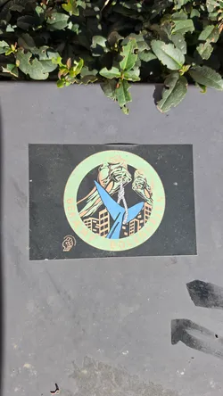 Street sticker A rectangular black sticker featuring a circular emblem at its center. Inside the mint-green ring, hands are depicted hoisting a blue shark by its tail with a rope, suspended above silhouettes of urban apartment blocks. The text 'BEKÆMP BOLIGHAJEN' curves around the lower part of the ring in light orange lettering. In the bottom-left corner sits a small Socialistisk Ungdomsfront (SUF) logo showing a clenched fist.