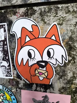 A die-cut sticker features an illustration of an orange and white fox holding a small envelope with a pink heart on it. The fox has large eyes with eyelashes. The fox is positioned in the center of the frame.