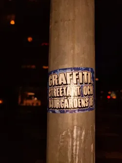 A sticker on a pole at night. The sticker has a blue background with white text. The text says "Graffiti, Street Art och Djurgårdens IF".