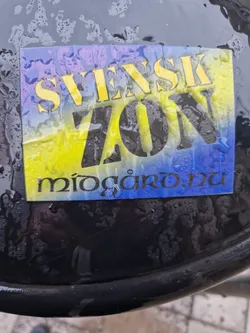 A sticker with the text "Svensk Zon" in yellow and black on a blue and yellow background. At the bottom, there is a website address: "miogaro.nu". The sticker appears to be affixed to a dark-colored surface that is wet.