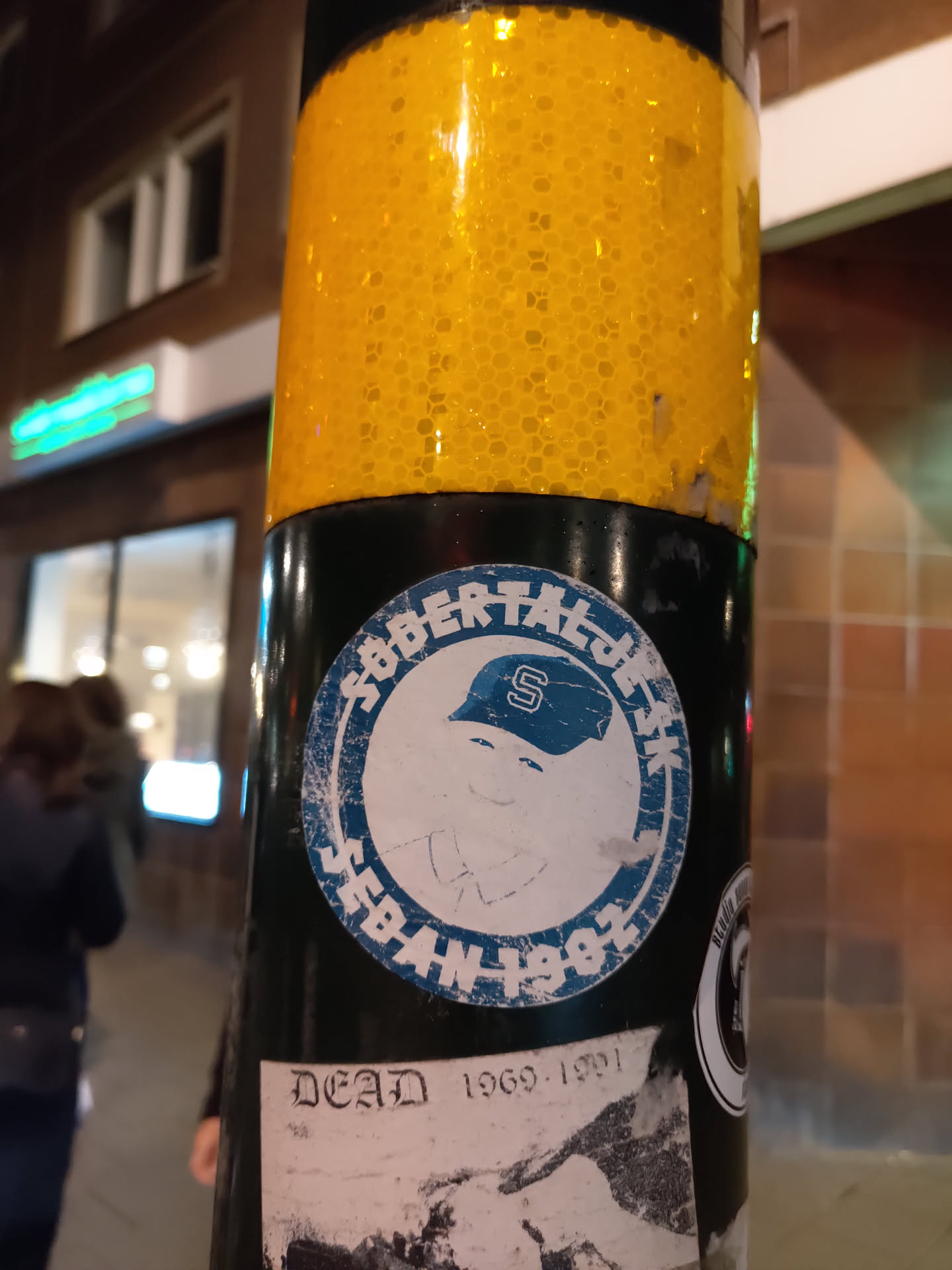 A worn sticker featuring a cartoon character wearing a baseball cap with the letter 'S'. The text 'Seban 1982' is visible within a circular design. Below this sticker, there is another sticker with 'Dead 1969-1991' written on it. 