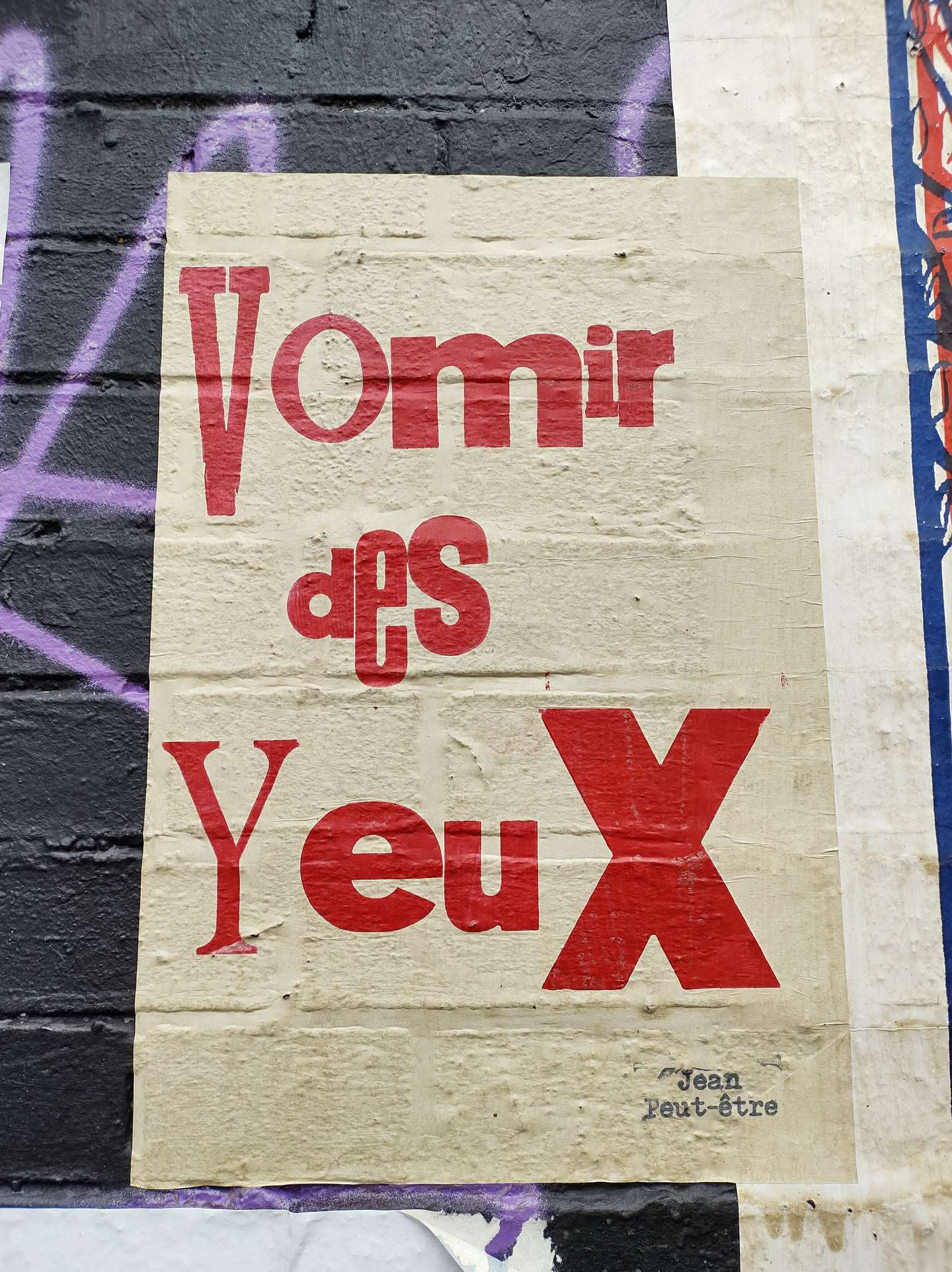 Street art poster with the text "Vomir des Yeux" in red and white. The text is in French and means "to vomit eyes". The poster is made of beige paper and has a rough texture. In the bottom right corner, there is a small text that says "Jean Peut-être", which could be the artist's name or signature.