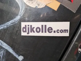 A rectangular sticker with a white background. The text 'djkolle.com' is printed in a dark purple sans-serif font. The sticker appears to be adhered to a dark, textured surface with some scuff marks and other stickers visible in the background.