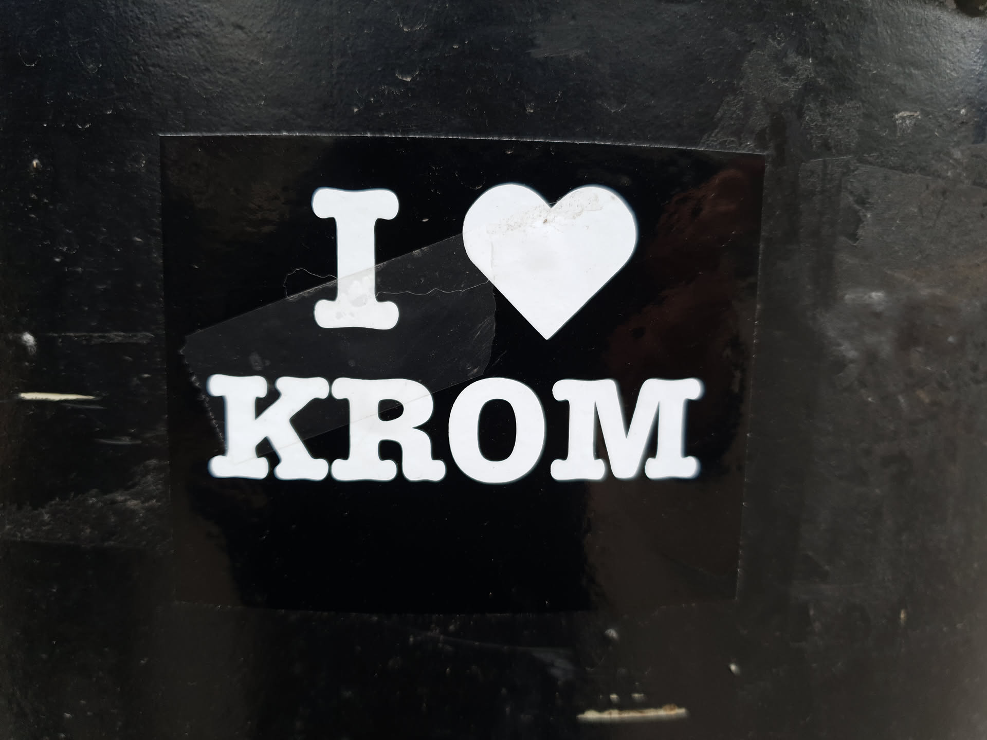 A black and white sticker with the text "I ♥ KROM" on it. The sticker is attached to a dark surface.