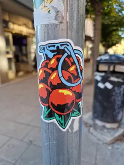 A sticker depicting a grenade-like object filled with red fruits, with green leaves at the bottom and a light blue handle. The style is reminiscent of traditional tattoo art.