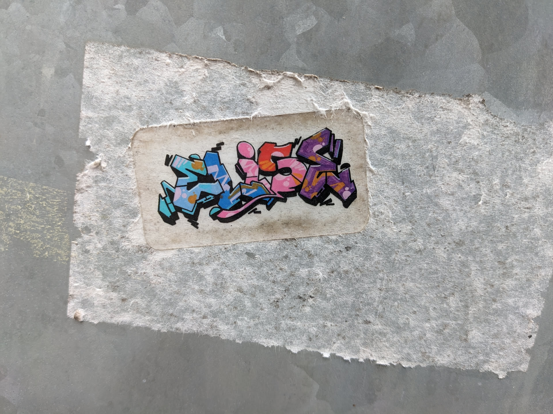 The sticker features graffiti-style lettering that appears to spell out the name 'ALON'. The letters are stylized with thick black outlines and filled with a gradient of colors. The first letter is predominantly blue with light blue and pink accents. The second letter is bright pink with orange highlights. The third letter is a mix of orange and pink with darker pink accents. The final letter is a deep purple with orange and yellow accents within its structure, and it has a shadow effect. The sticker itself is slightly aged and worn, with visible texture and some discoloration on its white background, attached to a rough, textured grey surface.