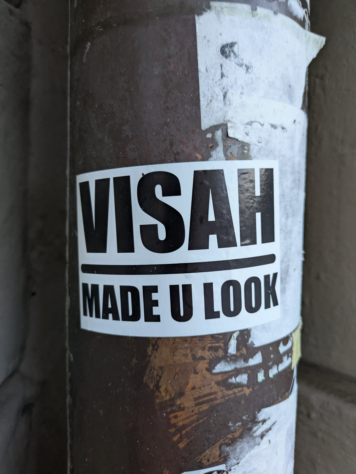 The sticker is white with bold black sans-serif lettering. The top line reads 'VISAH' in all caps. Below it, separated by a thin black line, is the phrase 'MADE U LOOK' in all caps. The sticker is applied to a weathered, cylindrical surface, likely a pole or pipe, which appears to be metal with rust and peeling paint underneath some torn white paper or remnants of other stickers.