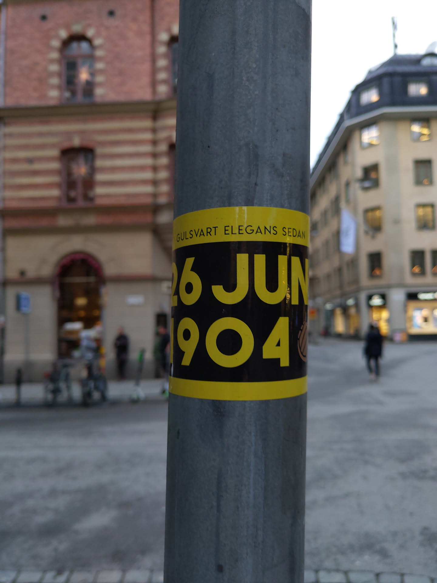 A black and yellow sticker affixed to a lamppost. The sticker features the text "GULSVART ELEGANS SEDAN" and the date "26 JUN 1904" prominently displayed in yellow on a black background. The context suggests a possible historical reference or event.