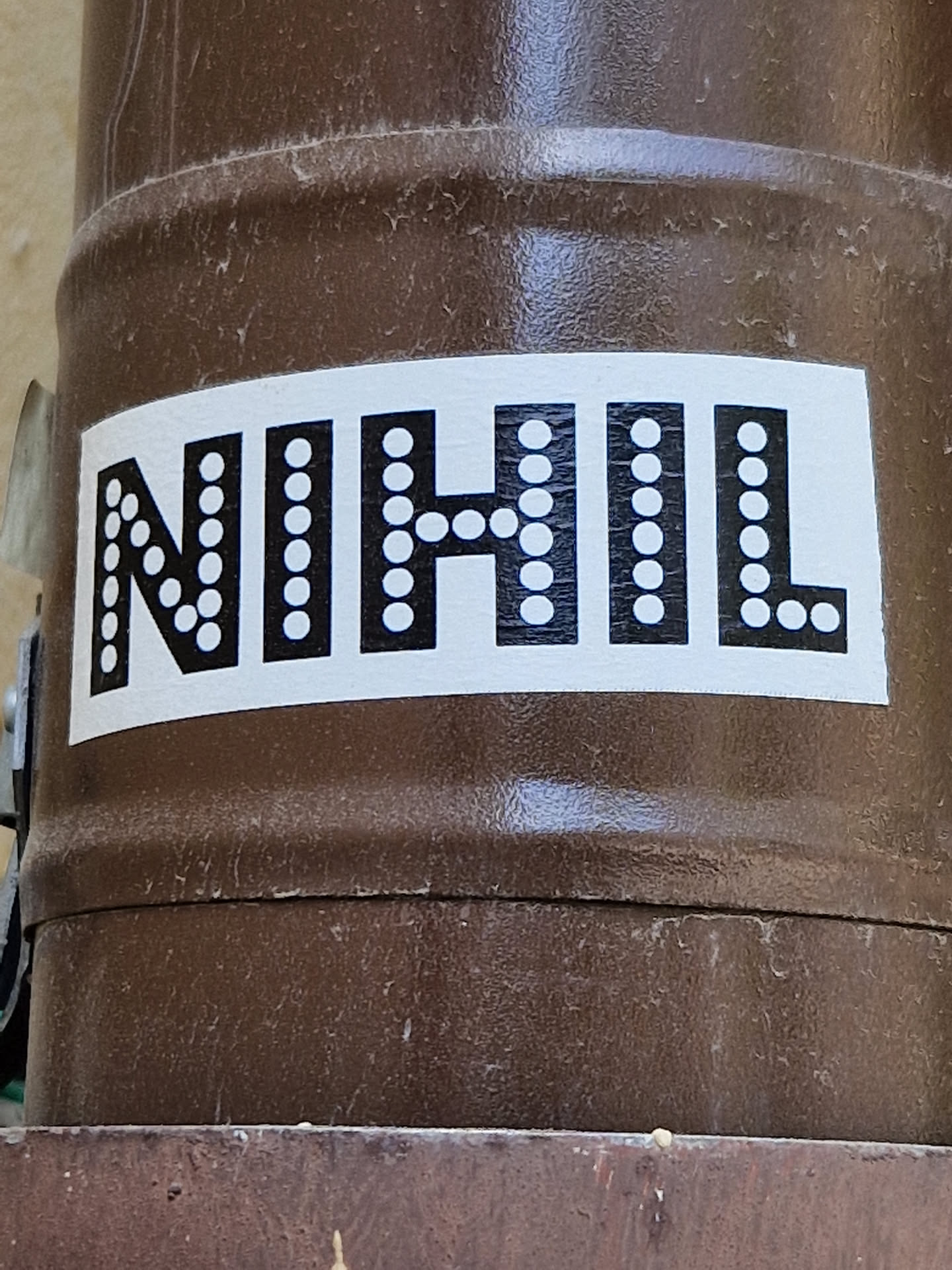 A rectangular sticker with the word NIHIL written in bold, uppercase letters.  Each letter is outlined with small white dots. The sticker is affixed to a brown cylindrical surface.
