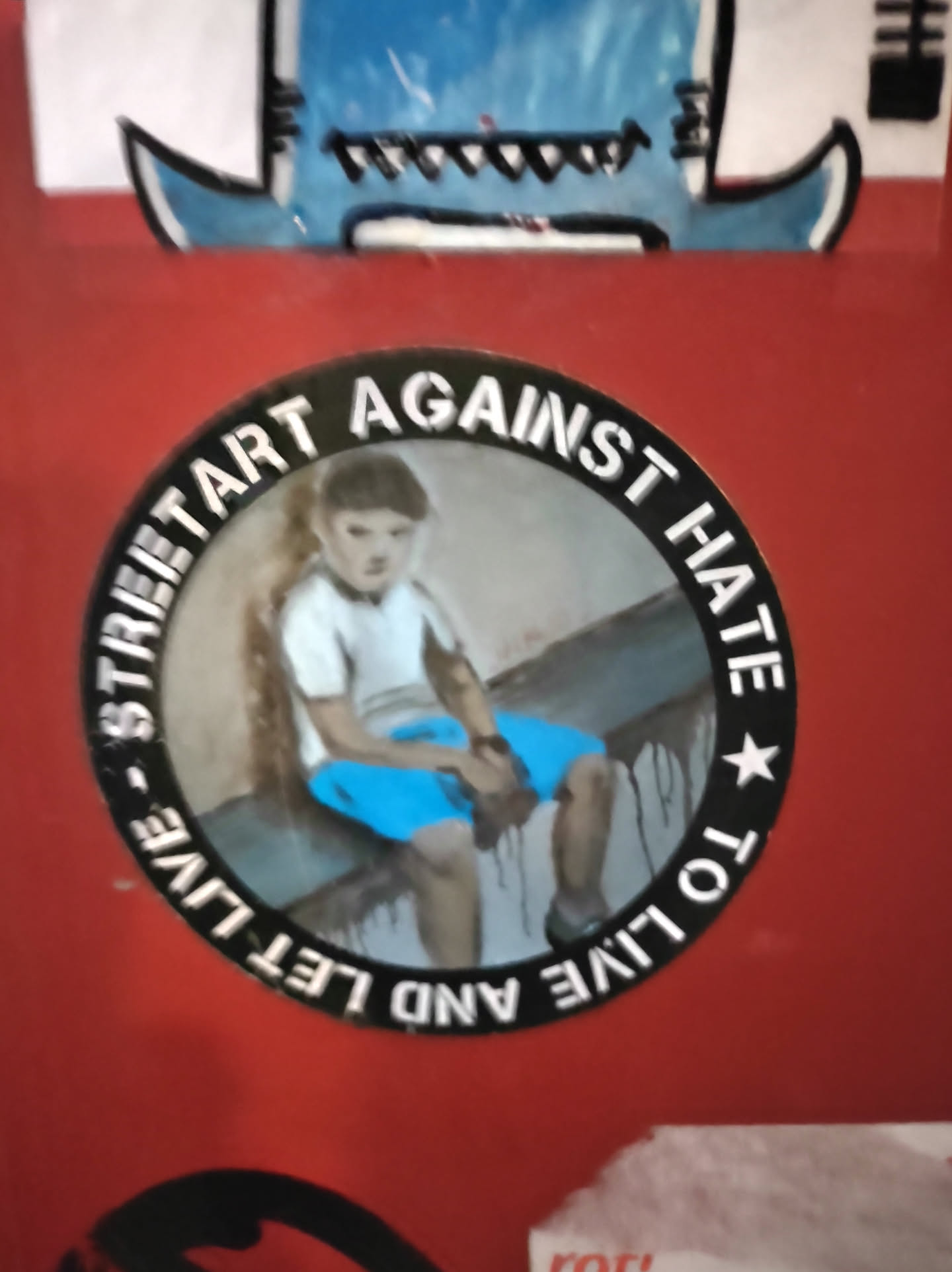 A round sticker with a picture of a child sitting on a wall. The text around the picture says "Street art against hate. To live and let live."
