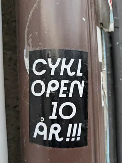 Black and white sticker on a brown pole.  The text on the sticker says "CYKL OPEN 10 ÅR!!!"