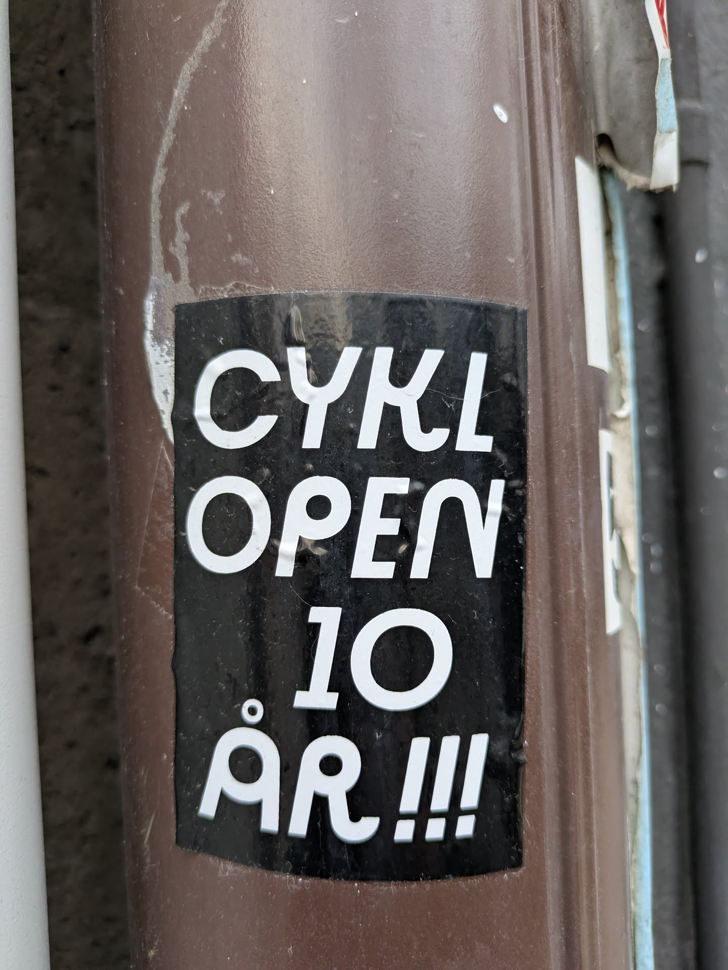 Black and white sticker on a brown pole.  The text on the sticker says "CYKL OPEN 10 ÅR!!!"