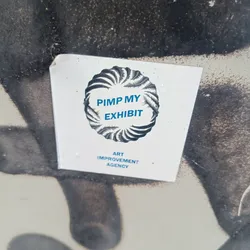 A sticker featuring a circular design with the text "Pimp My Exhibit" and the words "Art Improvement Agency" below. The sticker appears to be affixed to a wall or surface with some graffiti or markings.
