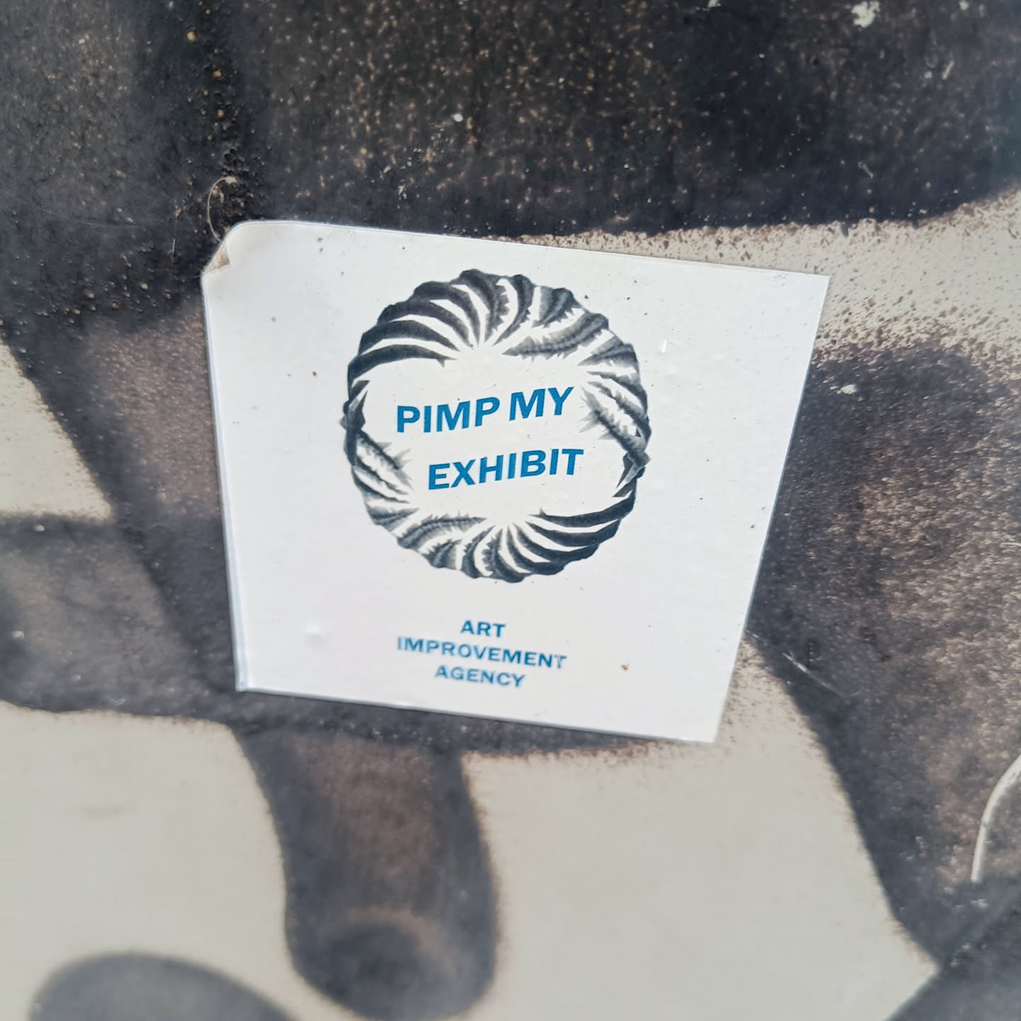 A sticker featuring a circular design with the text "Pimp My Exhibit" and the words "Art Improvement Agency" below. The sticker appears to be affixed to a wall or surface with some graffiti or markings.