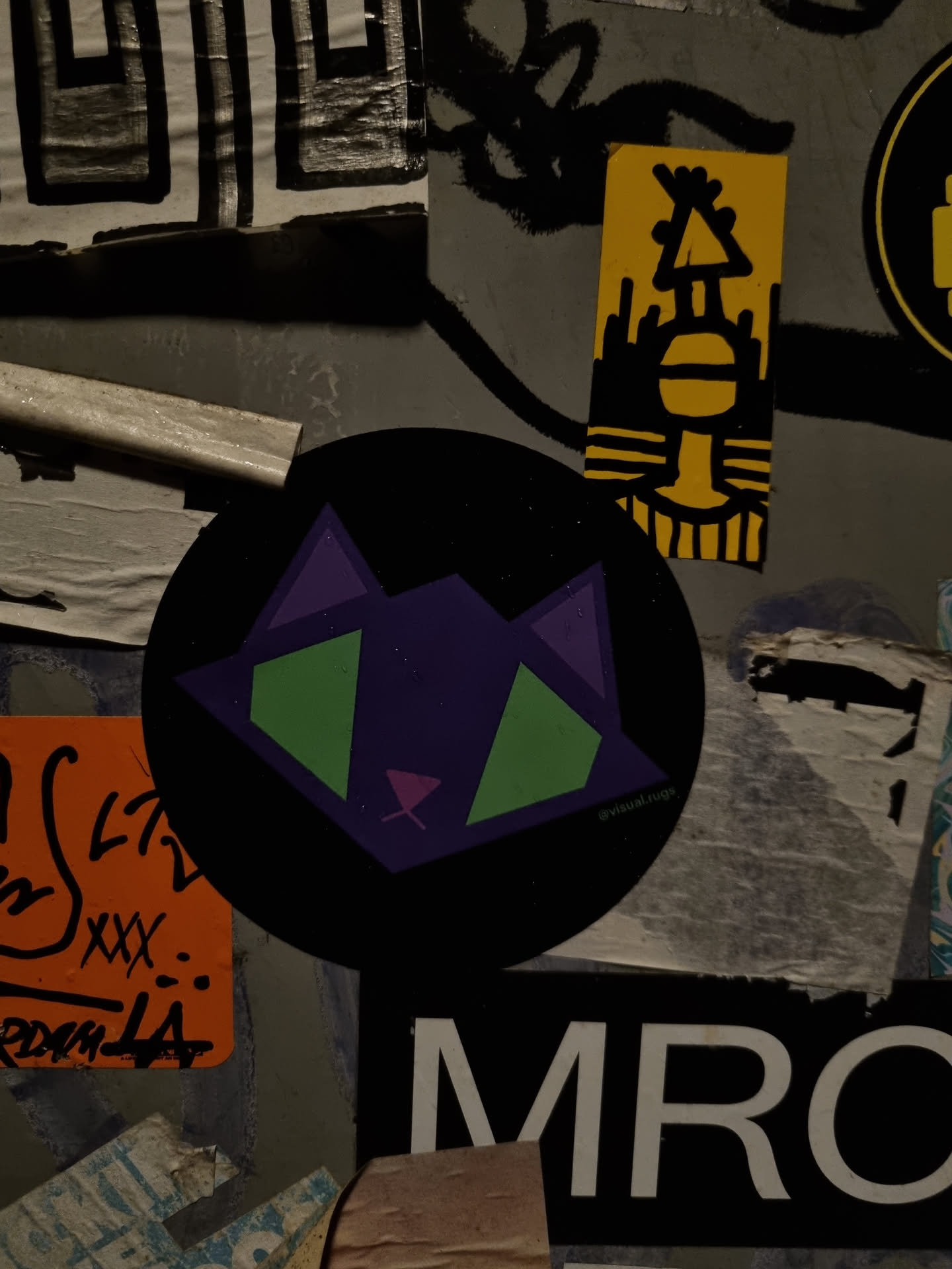 A round black sticker with a geometric design of a cat in purple, green, and pink. The artist's Instagram handle is @vivid.lugg.