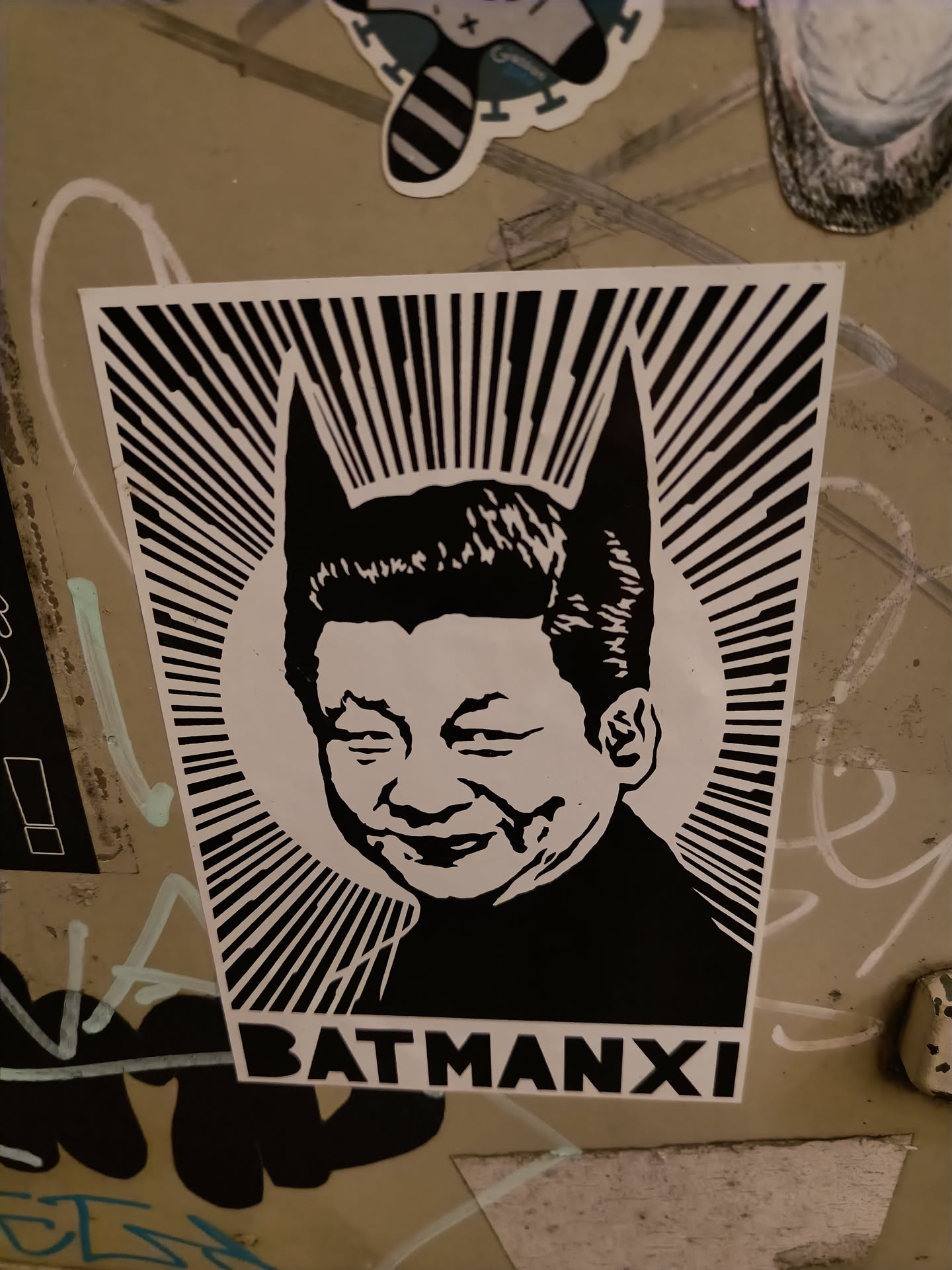A black and white sticker depicting Xi Jinping with bat ears, resembling Batman. The sticker has a bold, graphic style with radiating lines behind the portrait and the text 'BATMANXI' at the bottom.