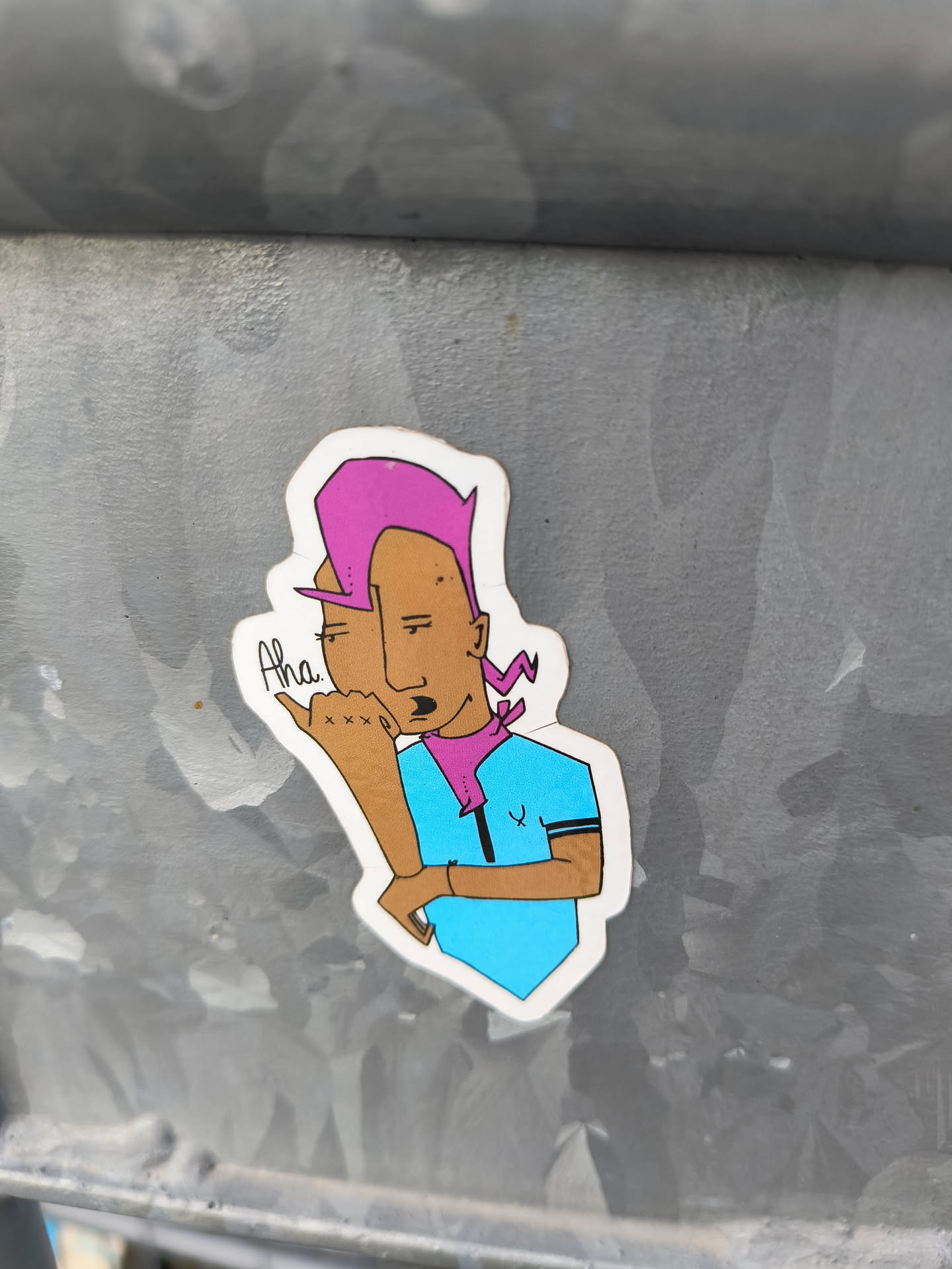 This sticker features a cartoon-like illustration of a person with pink hair, a purple bandana, and a light blue shirt. The character has a playful expression and appears to be making a gesture. The art style is simple yet expressive. The sticker is affixed to a metallic surface.