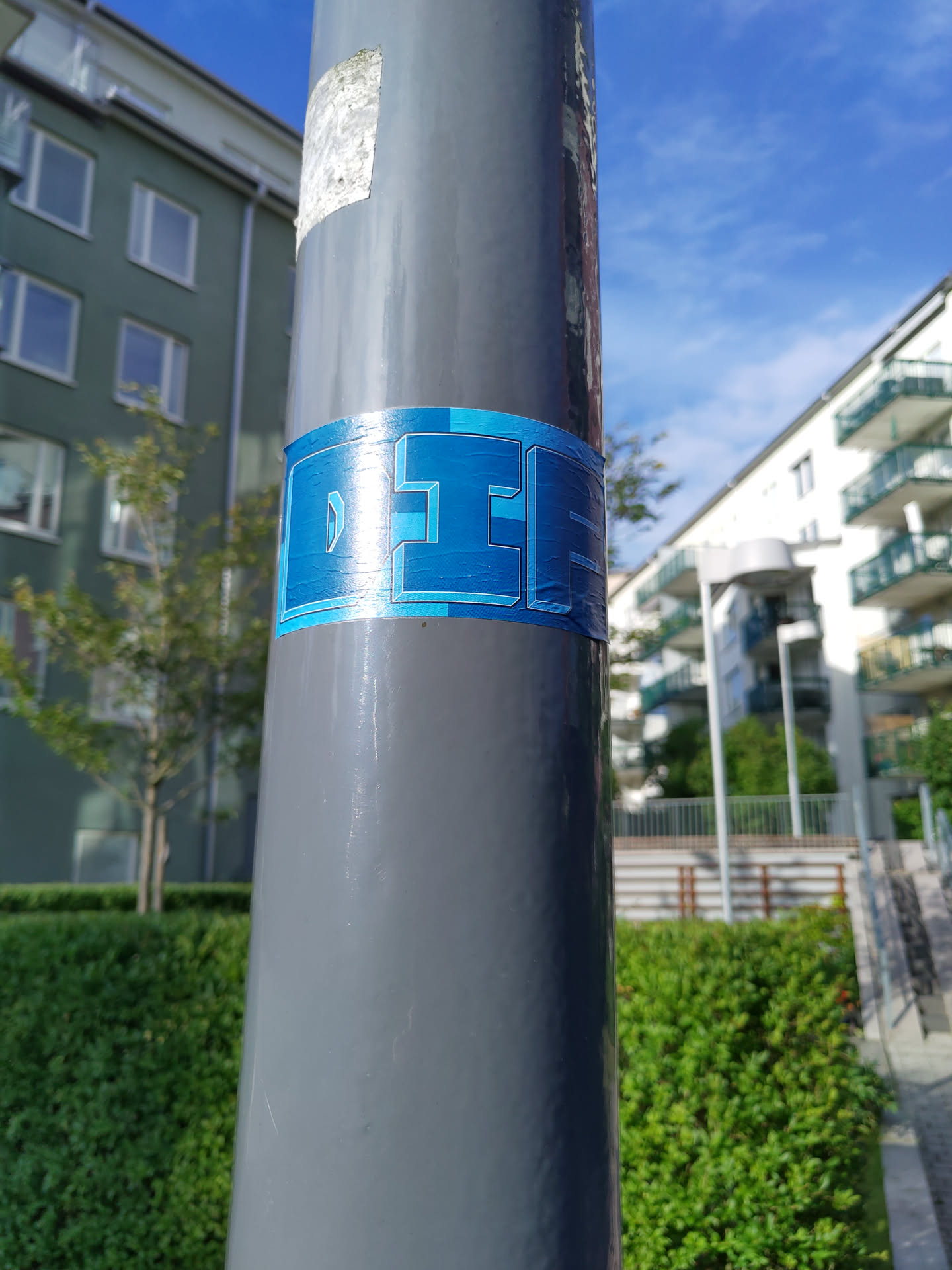 A close-up shot of a blue and gray sticker on a gray pole. The sticker features a stylized text that seems to be an acronym or a logo. The background is blurry, showing residential buildings and greenery. The overall impression is one of urban minimalism.