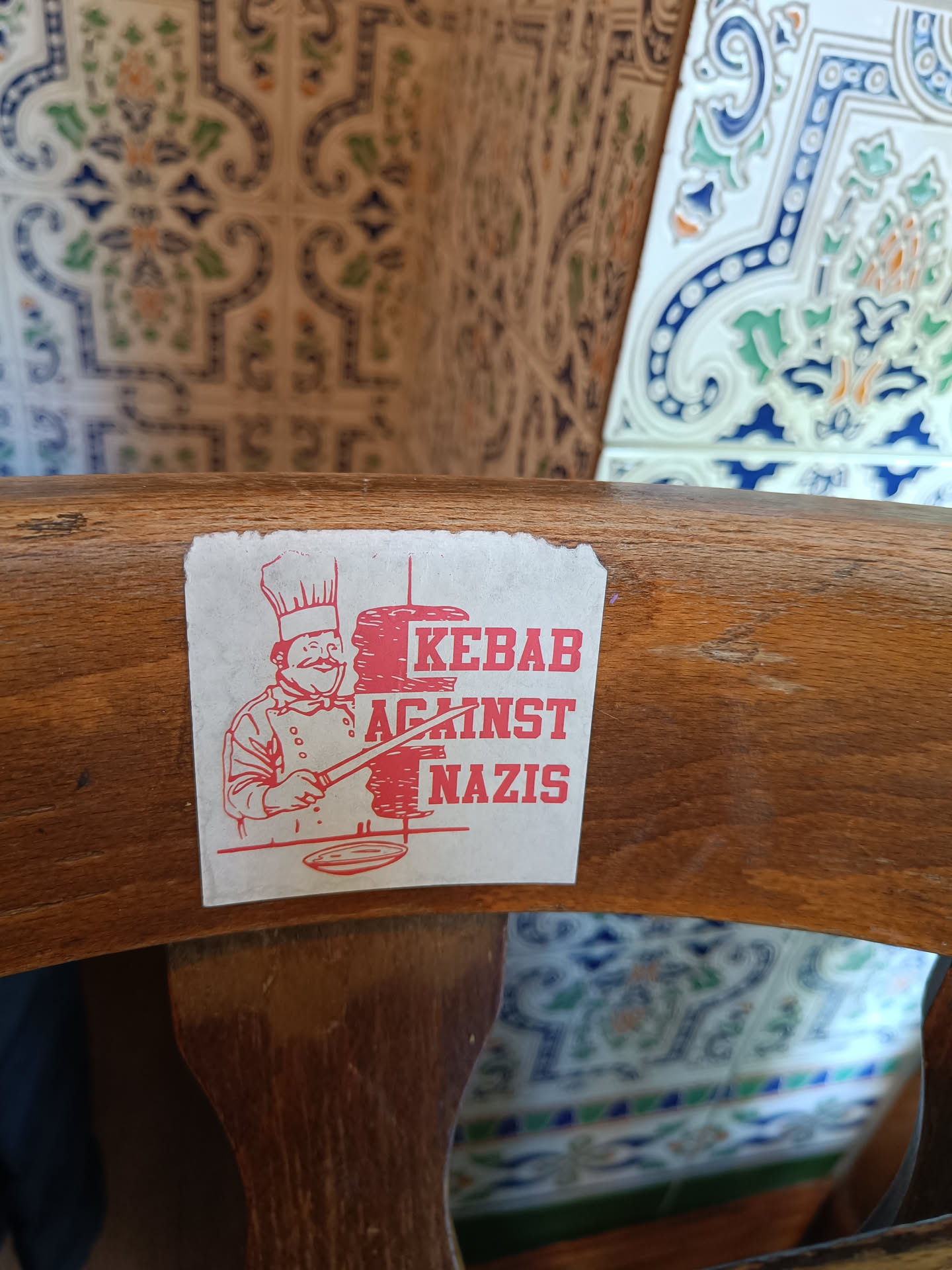 A sticker featuring a chef preparing a kebab with the text "KEBAB AGAINST NAZIS". The sticker is affixed to a wooden chair.