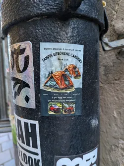 A vertical rectangular sticker pasted onto a textured black metal post. The upper part features an illustration of two orange camping tents pitched in front of a misty pine forest, framed by the text 'Explore, Discover & Love Our Nature' and 'TAMPIN GEBORENE CAMPERS Since 2025'. The lower half has a dark background reading 'GAMES | MUSIC | FUN | ADVENTURE' above the inspirational quote 'If you truly love nature, you will find beauty everywhere!'. Below the quote are two small color photos showing off-road vehicles (a red pickup truck and an orange 4x4) outdoors.