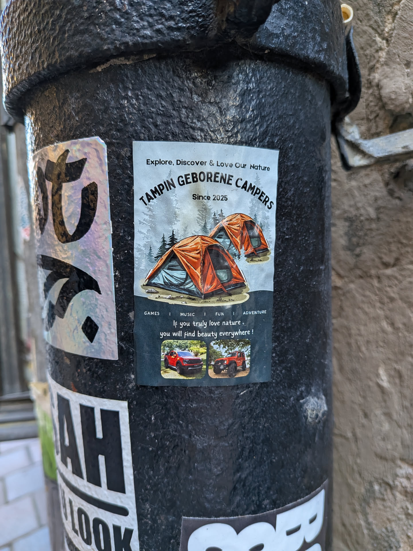 A vertical rectangular sticker pasted onto a textured black metal post. The upper part features an illustration of two orange camping tents pitched in front of a misty pine forest, framed by the text 'Explore, Discover & Love Our Nature' and 'TAMPIN GEBORENE CAMPERS Since 2025'. The lower half has a dark background reading 'GAMES | MUSIC | FUN | ADVENTURE' above the inspirational quote 'If you truly love nature, you will find beauty everywhere!'. Below the quote are two small color photos showing off-road vehicles (a red pickup truck and an orange 4x4) outdoors.