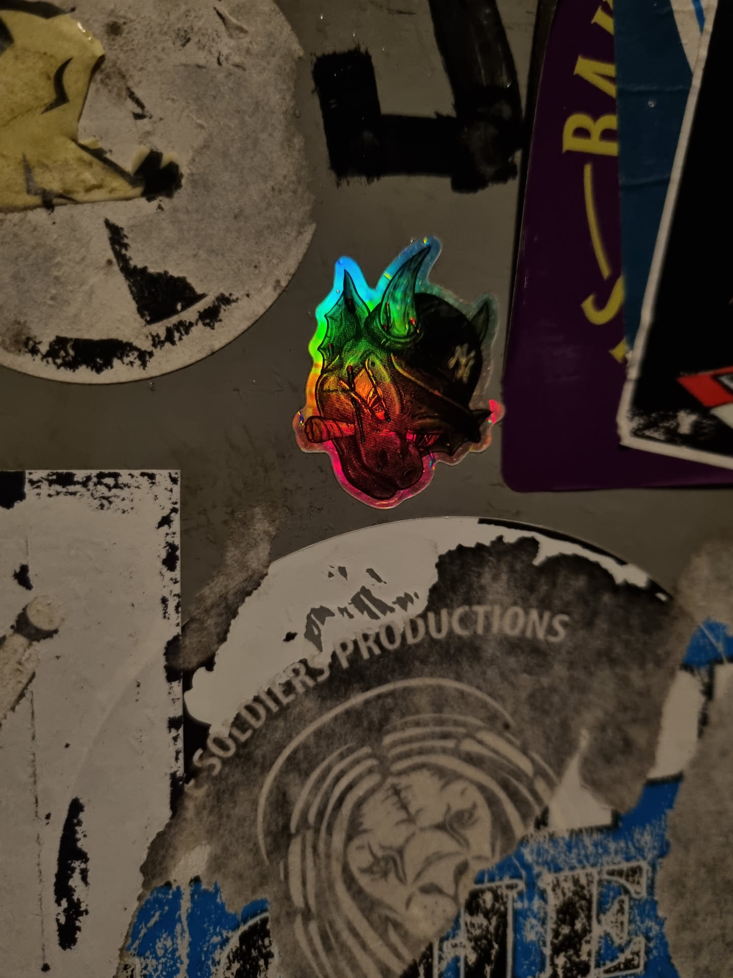 A small, holographic sticker featuring a demonic-looking head wearing a pirate hat. The sticker has a rainbow effect and is affixed to a wall among other stickers and graffiti.  There is text on a nearby sticker that says "Soldiers Productions."
