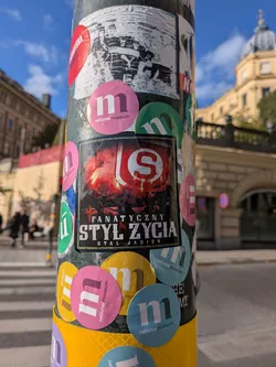 Street sticker A rectangular sticker placed on a pole amidst several round Nationalmuseum stickers. The central sticker features a dramatic image of stadium supporters holding glowing red pyrotechnics and flares in the background. In the upper right overlay is a red and white emblem with the letter 'S'. Bold white and stylized typography at the bottom reads 'FANATYCZNY STYL ŻYCIA' with 'STAL JASIEŃ' printed underneath.