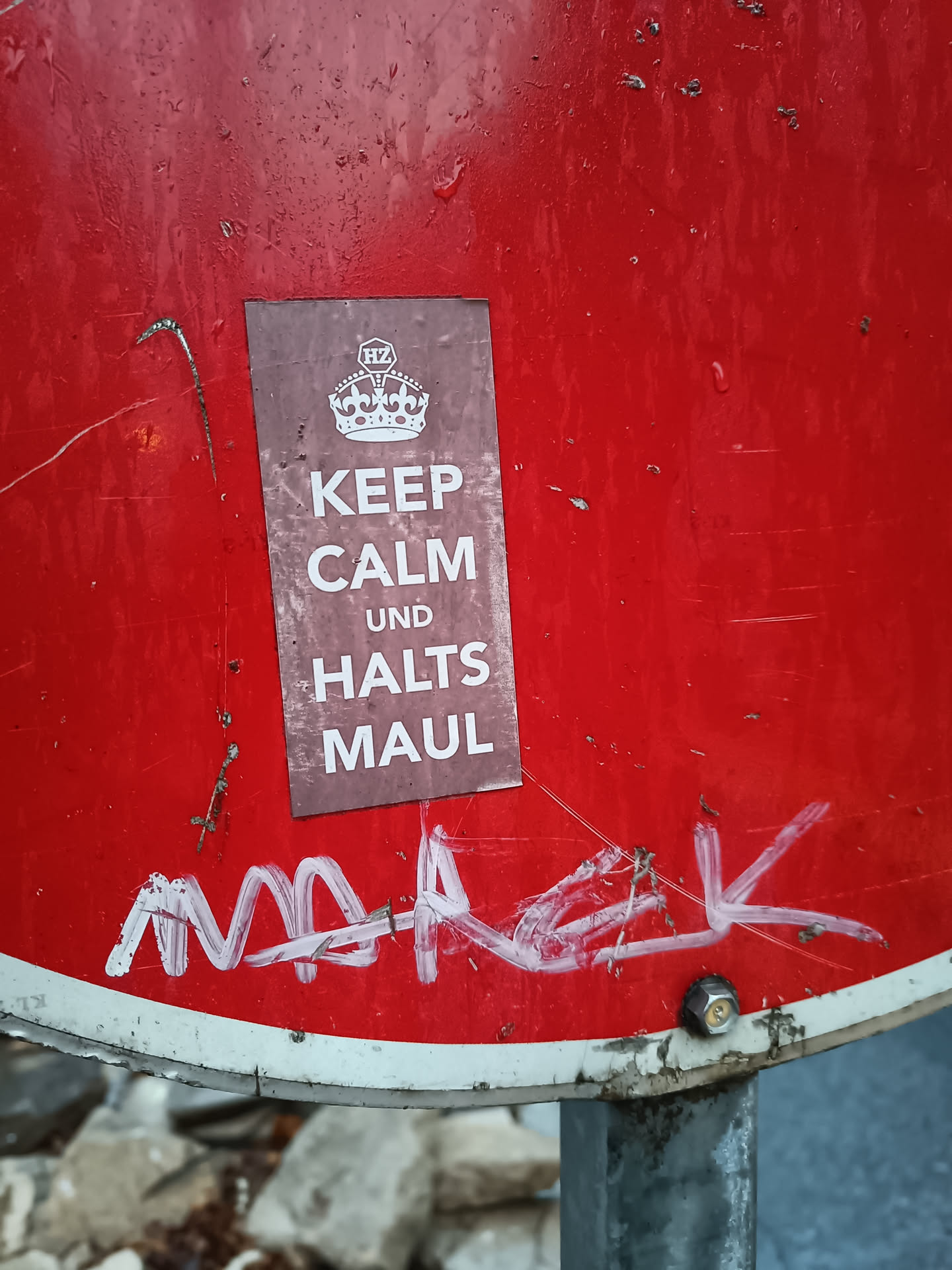 A sticker on a red sign that says "Keep Calm und Halts Maul" with a crown on the top.  There is also some white graffiti on the sign.