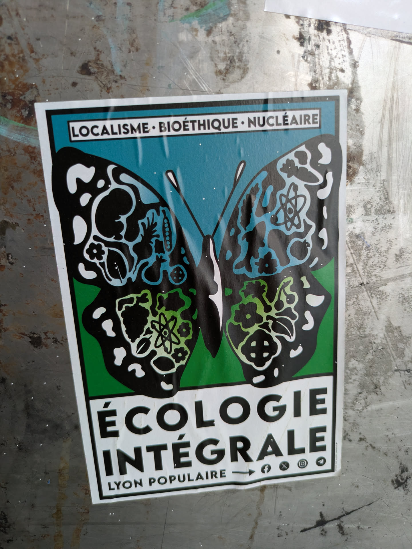 The sticker is a rectangular piece of graphic art, predominantly featuring a black butterfly with intricate white, blue, and green patterns on its wings, set against a split background of teal on top and green on the bottom. The upper wings display abstract organic shapes, including what appears to be a fetus-like figure and other natural elements. The lower wings contain symbols such as an atom and flowers, suggesting a blend of scientific and natural themes. Above the butterfly, the text "LOCALISME • BIOÉTHIQUE • NUCLÉAIRE" is prominently displayed. Below the butterfly, in large bold letters, it reads "ÉCOLOGIE INTÉGRALE". At the very bottom, the words "LYON POPULAIRE" are visible, alongside icons indicating social media presence on Facebook, X (formerly Twitter), Instagram, and Telegram.
