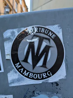 A circular sticker featuring a prominent black and white design. The central element is a stylized monogram of the letters 'TM', with the 'T' positioned above and slightly behind the 'M'. Both letters have a sharp, angular serif style with a white outline and black fill. Behind the monogram, there are concentric circles in varying shades of grey, creating a subtle target or ripple effect. Along the top curved edge of the circle, the word 'TRIBUNE' is written in white serif capital letters with a black outline. Similarly, along the bottom curved edge, the word 'MAMBOURG' is written in the same style. The sticker has a glossy finish, showing some reflections, and is adhered to a textured grey surface, with visible remnants of adhesive or other stickers around its edges.
