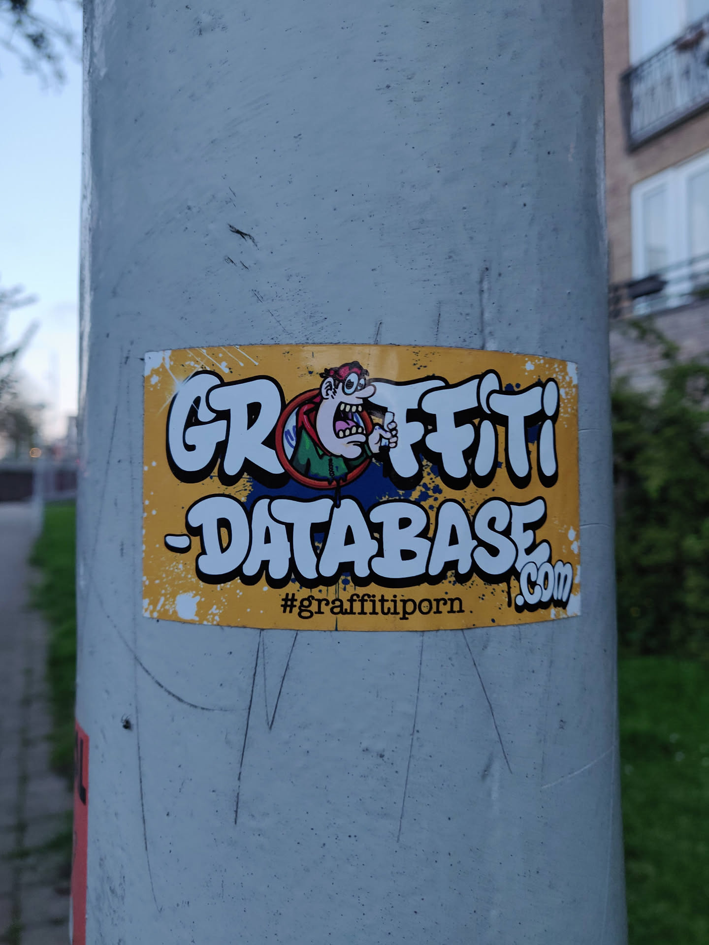 A sticker featuring a cartoon character and the text "Graffiti -Database.com" with the hashtag #graffitiporn at the bottom. The sticker is yellow with black lettering and is affixed to a gray pole.