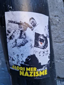 A sticker depicting a woman aiming a rifle, with three other figures in the background.  A black flag with a symbol is also present. The text 'ALDRI MER NAZISME' is visible at the bottom, along with 'ANTIFA.TK'. The image appears to be a protest against Nazism.