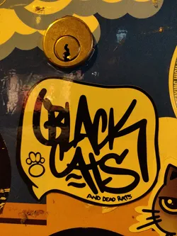 A yellow sticker with black graffiti-style text that says "Black Cats" with a small paw print and the words "and Dead Rats" underneath. The sticker is speech bubble-shaped and is affixed to a surface with other stickers.