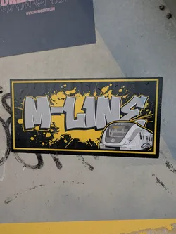 A sticker featuring graffiti-style lettering of 'M-Line' with a train illustration. The design incorporates a yellow and black color scheme with paint splatters.