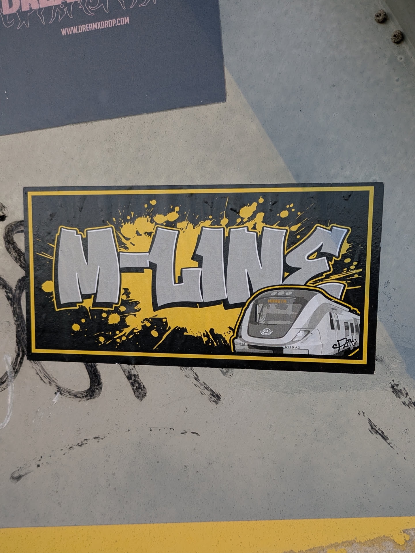 A sticker featuring graffiti-style lettering of 'M-Line' with a train illustration. The design incorporates a yellow and black color scheme with paint splatters.