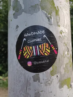 Black round sticker with the text 'HANDMADE CLOTHES' in the center. It also contains an illustration of yarn and knitting needles. At the bottom, there is an Instagram handle: @paolakttal