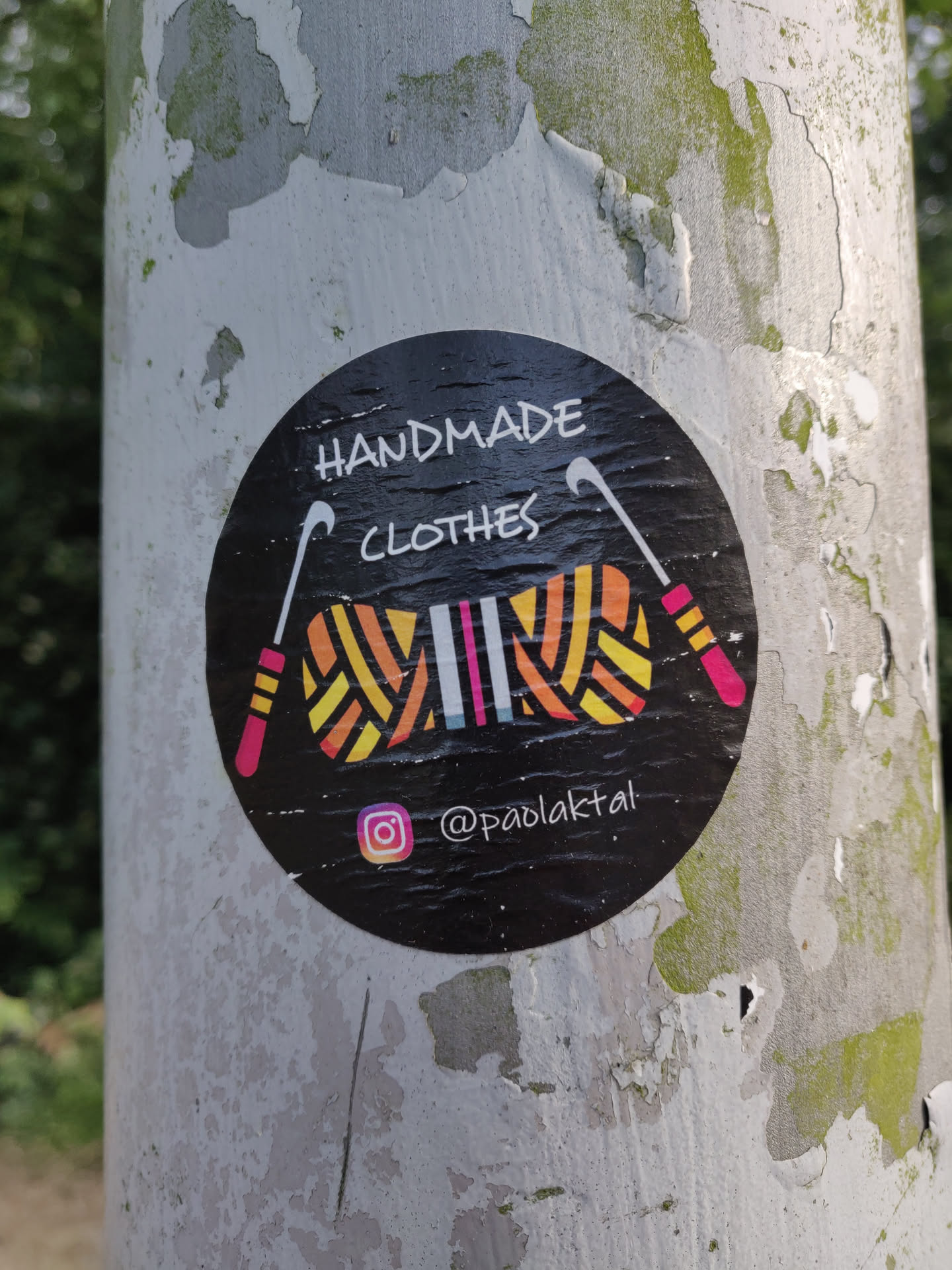Black round sticker with the text 'HANDMADE CLOTHES' in the center. It also contains an illustration of yarn and knitting needles. At the bottom, there is an Instagram handle: @paolakttal