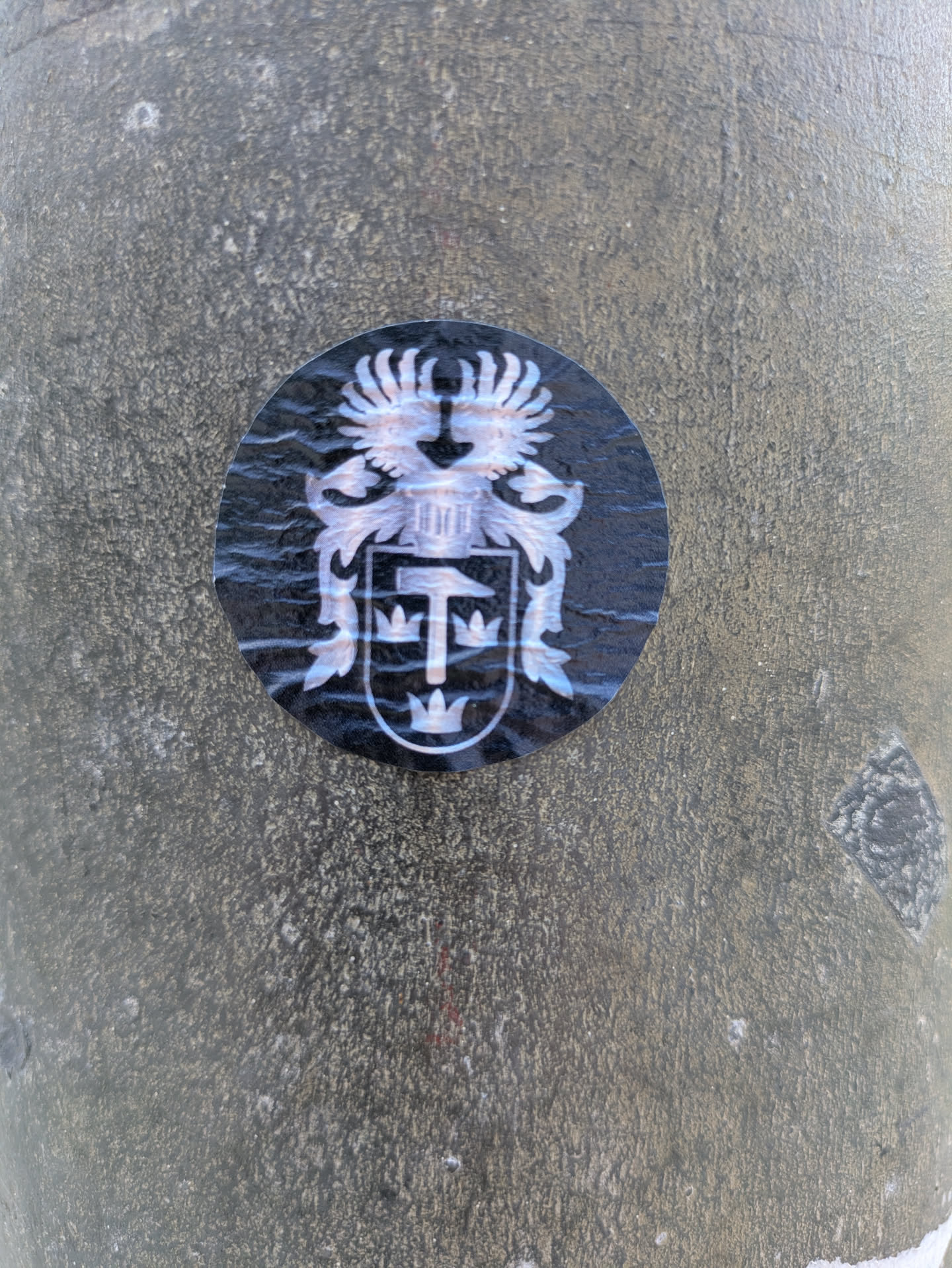 A close-up of a circular sticker featuring the coat of arms of Kiruna, Sweden. The emblem is rendered in white or silver on a black background. It depicts a heraldic shield with a central mining hammer and pickaxe, symbolizing the city's primary industry, surrounded by three crowns (two above, one below), representing Sweden. Above the shield, there is a helmet with a crest resembling stylized wings or plumes. The sticker is affixed to a textured, possibly concrete or metal, surface with a greyish-brown color.