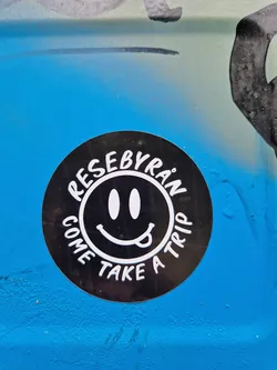 Round black sticker with a smiley face and the text "Rese by Ran" written around the smiley. The text "Come take a trip" is written below the smiley face.