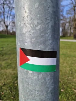 A sticker depicting the flag of Palestine is affixed to a metallic pole outdoors. The background shows a grassy area with trees in the distance. The flag is clearly visible and appears to be made of a vinyl or similar material.