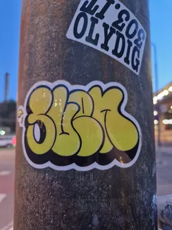 Close-up of a yellow and black sticker with graffiti-style lettering. The sticker is affixed to a gray pole. The lettering is stylized and bubbly.  There are other stickers visible on the pole but this is the main focus.