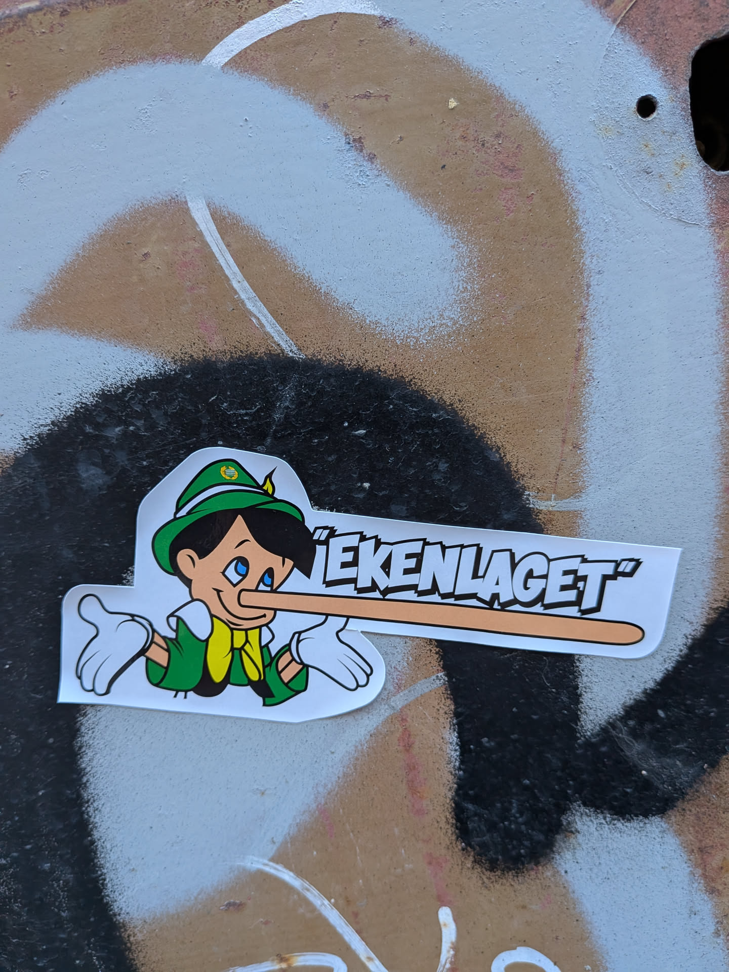 The sticker features a cartoon character resembling Pinocchio. He is depicted wearing a green hat with a yellow band and a white feather, a green jacket, a yellow scarf, and white gloves. His signature long nose is prominently extended, suggesting he is telling a lie. The character's eyes are blue and he has a slight smile. The sticker has a white border and is placed on a background with graffiti.
