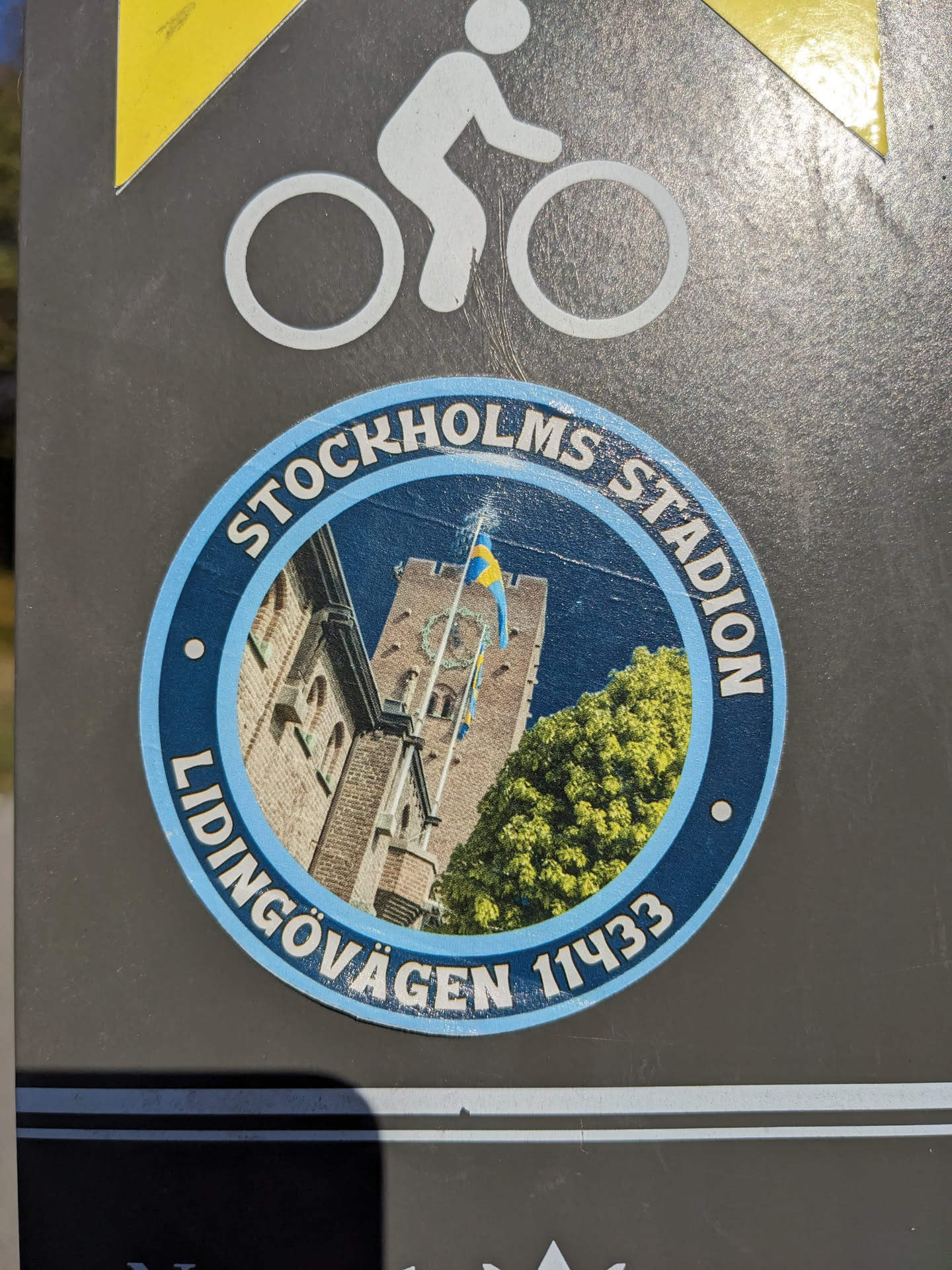 Round sticker featuring a picture of a building with a flag, and text around it that says "Stockholms Stadion" and "Lidingövgen 11433".
