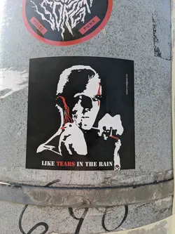 The sticker is predominantly black and white, featuring a stylized portrait of an androgynous figure with striking red accents on the eyes, mimicking tears. In the figure's hands, a white dove is held, and the figure appears to be holding a lit cigarette with red glowing tip. The background is solid black. White text is printed at the bottom of the sticker, and a small circular logo with a red 'R' inside it is also present. The overall aesthetic is stark and evocative.