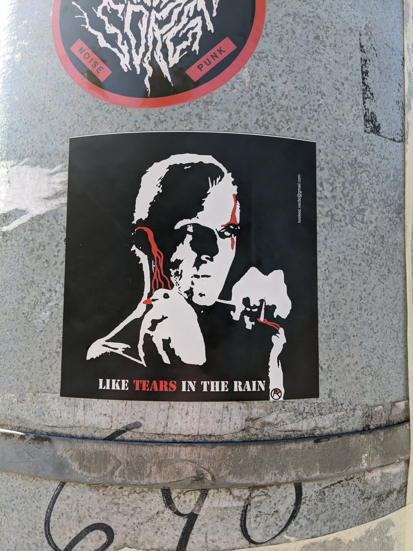 The sticker is predominantly black and white, featuring a stylized portrait of an androgynous figure with striking red accents on the eyes, mimicking tears. In the figure's hands, a white dove is held, and the figure appears to be holding a lit cigarette with red glowing tip. The background is solid black. White text is printed at the bottom of the sticker, and a small circular logo with a red 'R' inside it is also present. The overall aesthetic is stark and evocative.